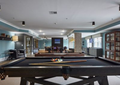 The game room at The Winston at Lyndhurst Apartments featuring a pool table in the foreground with billiard cues and balls, a shuffleboard table, seating, and a mounted TV in the background.