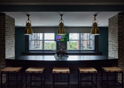 The community lounge at The Winston at Lyndhurst Apartments featuring an island with wooden barstools, pendant lighting, and large windows offering natural light and views of the surrounding apartment complex. A mounted TV is displayed in the background.